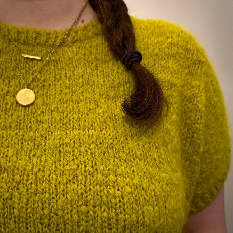 A close-up shot of a fluffy, hand-knit sweater. The sweater is chartreuse in color and has short sleeves. It appears soft to the touch.