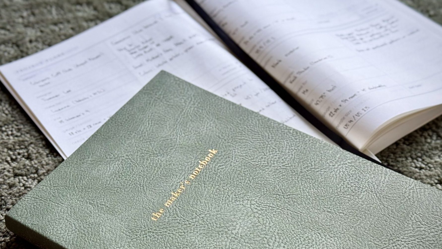 Two notebooks are photographed at an angle, one closed and resting on top of the other, which is opened to a page with blurred writing. The closed notebook is sage green and has “The Maker’s Notebook” printed in gilded letters on the cover.